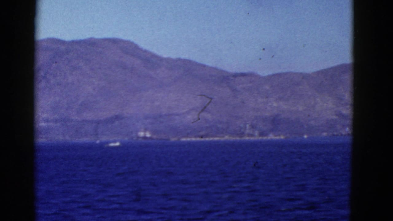 Hilly shoreline across blue water with a small vessel near the coast.