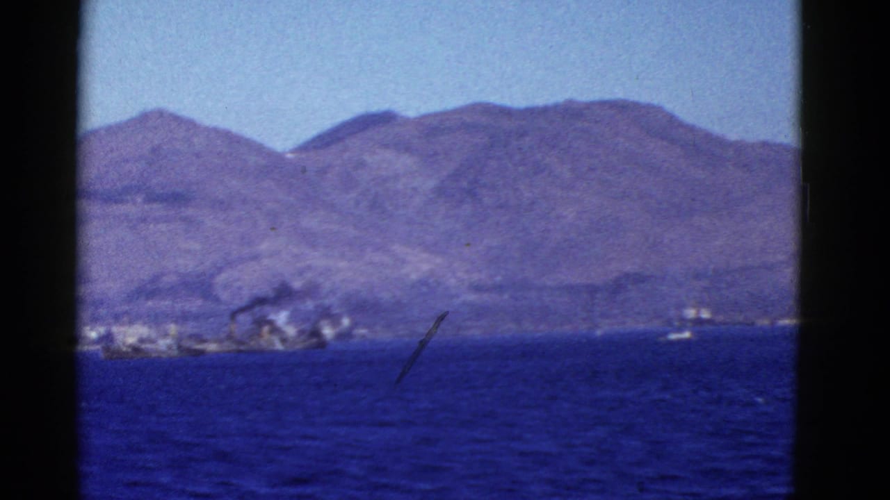 1950s 8mm view from San Francisco showing blue water, rugged mountains, and a distant smoky shoreline vessel or waterfront area.