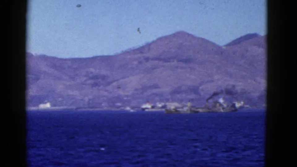1950s San Francisco coastal home-movie frame with blue water, a broad mountain ridge, and smoky activity near the far shore.