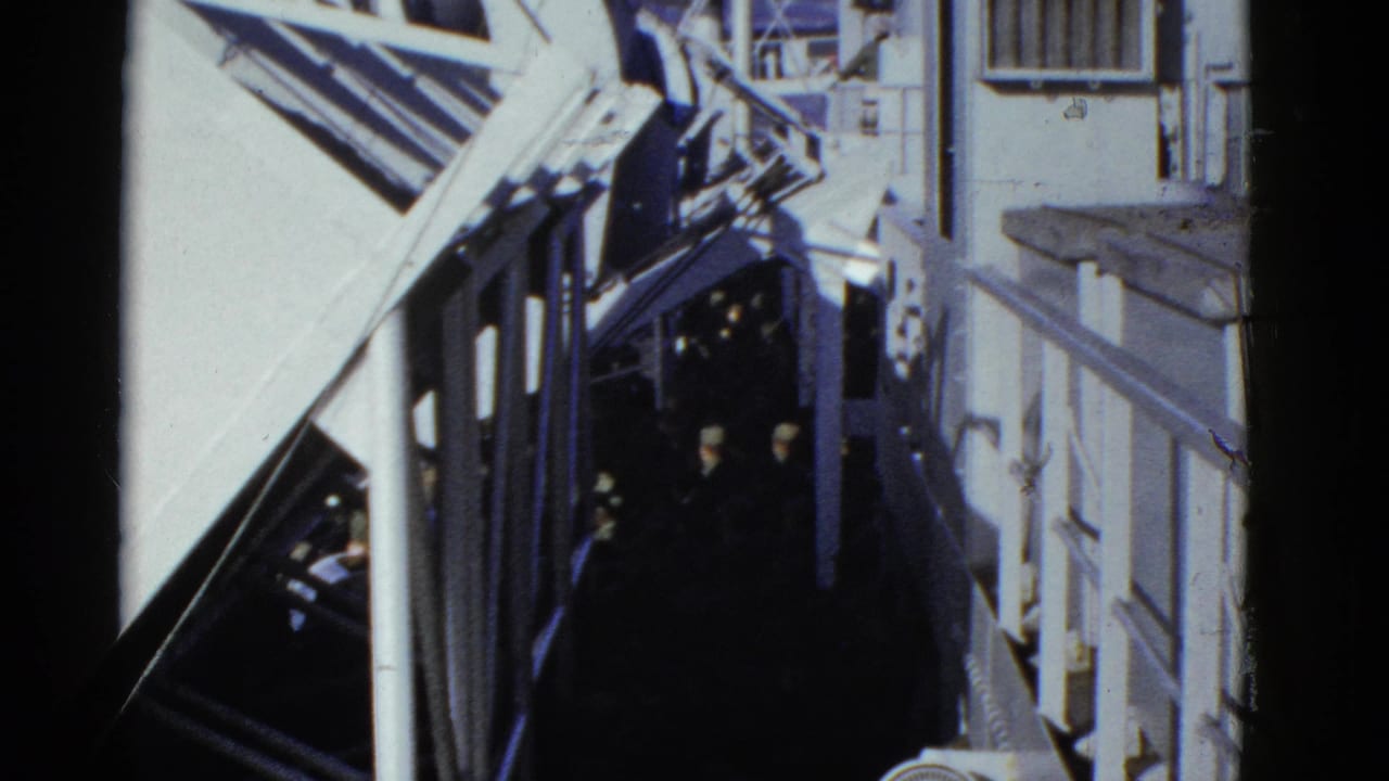 People in dark uniforms continue along a ship passage beside white railings and beams.