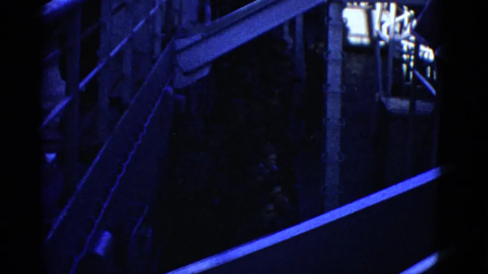 Blue-shifted 8mm frame of a dim shipboard corridor or lower passage with railings, stairs, and dark-uniformed figures.