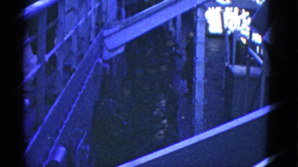 1952 home-movie frame showing a dim blue-toned ship passage with railings, posts, and partially visible uniformed people.