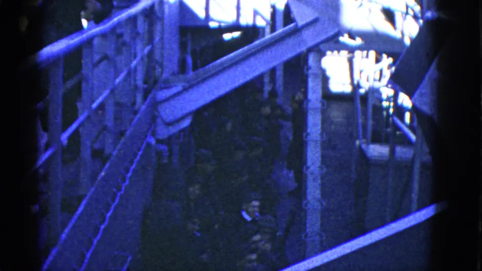 Blue-toned 8mm frame of a San Francisco dock area, with a raised walkway, railings and people partly obscured in shadow.