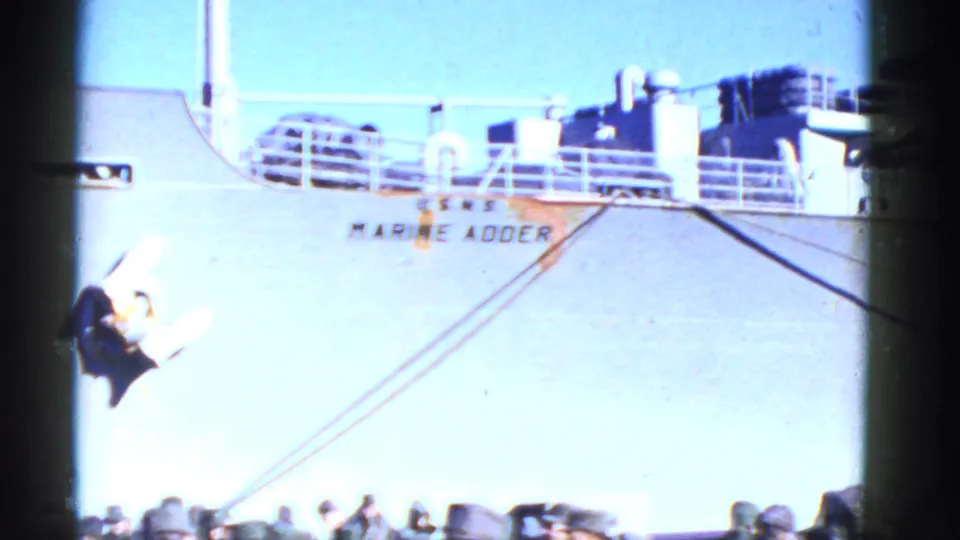 1950s San Francisco seaport frame showing a large ship side with painted name, deck railing, ropes and dockside people below.