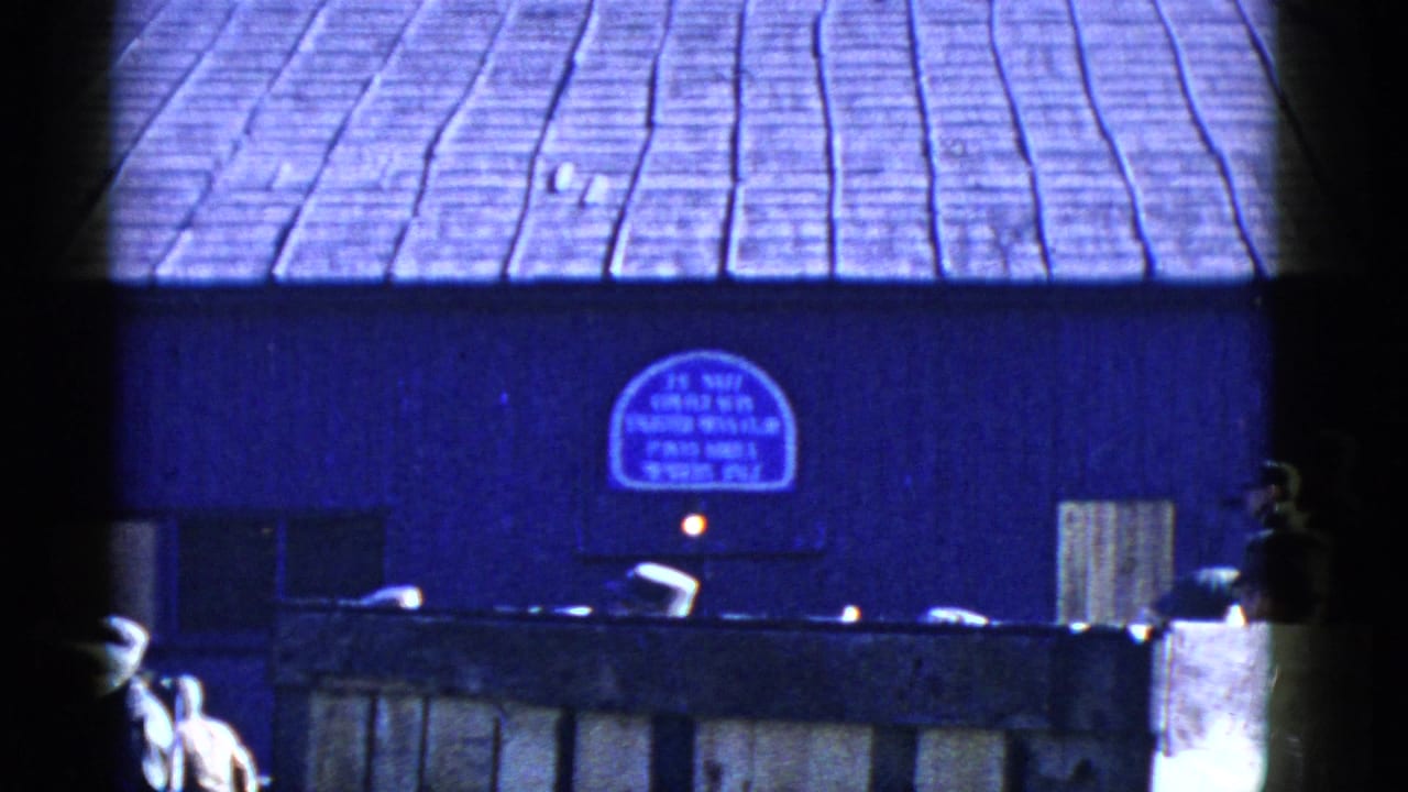 1950s San Francisco home-movie frame showing uniformed men near a wooden fence and blue building with a partially legible arched sign.