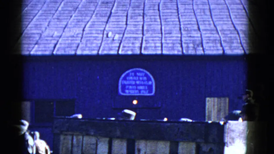 1950s San Francisco home-movie frame showing uniformed men near a wooden fence and blue building with a partially legible arched sign.