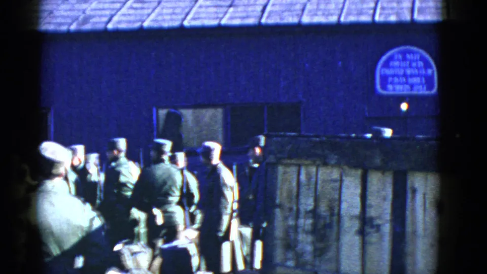 Several uniformed service members stand or walk near a wooden fence, with the blue building and arched wall sign behind them.