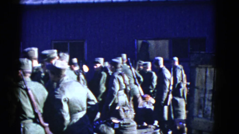 A larger group of uniformed men, including rifle carriers, gathers near the fence and blue building in this 1950s San Francisco frame.