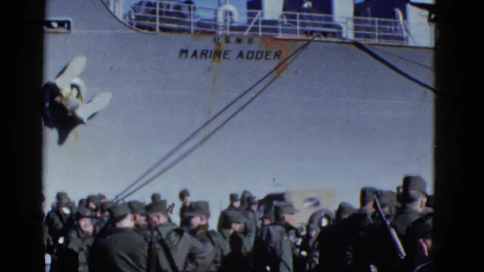 Military men stand in a crowded boarding area beneath the visible Marine Adder name on a large ship.
