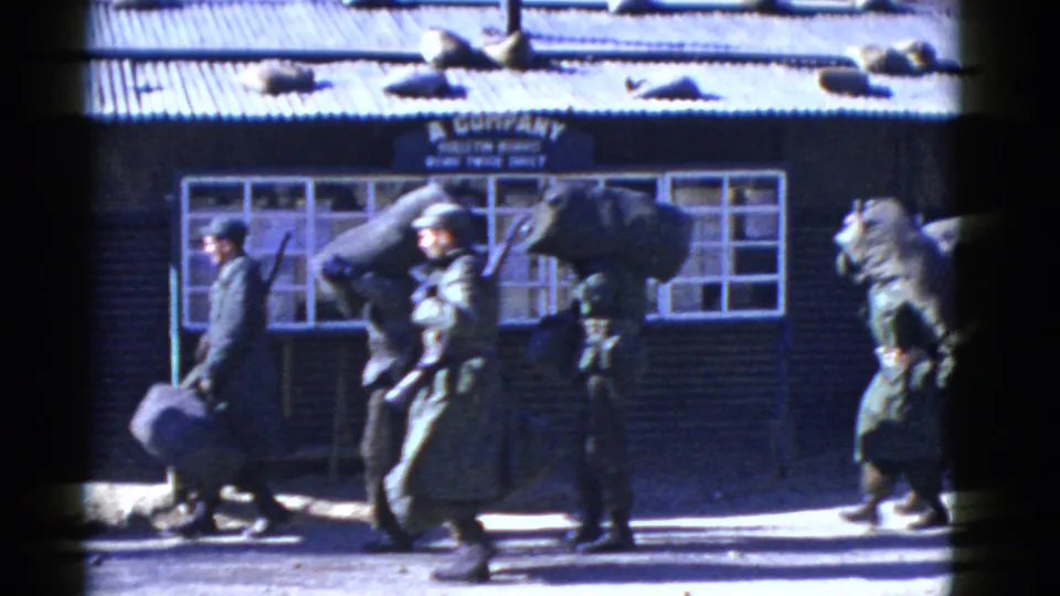 1950s military personnel carry duffel bags and packs past the A Company building in bright sun.