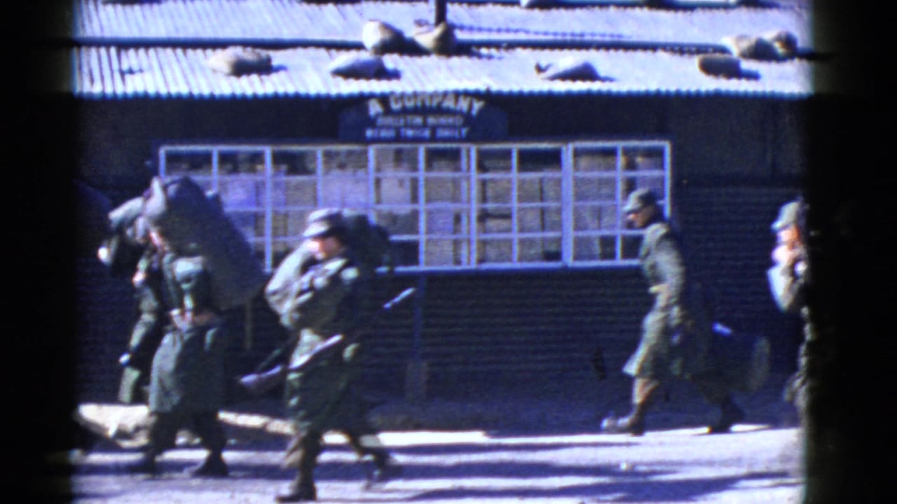 Uniformed soldiers, some carrying rifles and bulky packs, walk across the frame outside a barracks-like A Company building in the 1950s clip.