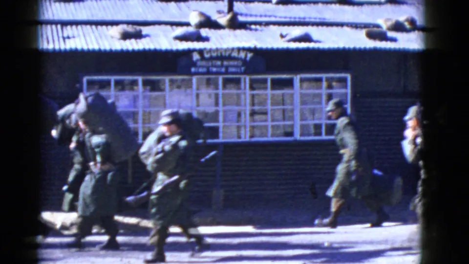 Uniformed soldiers, some carrying rifles and bulky packs, walk across the frame outside a barracks-like A Company building in the 1950s clip.