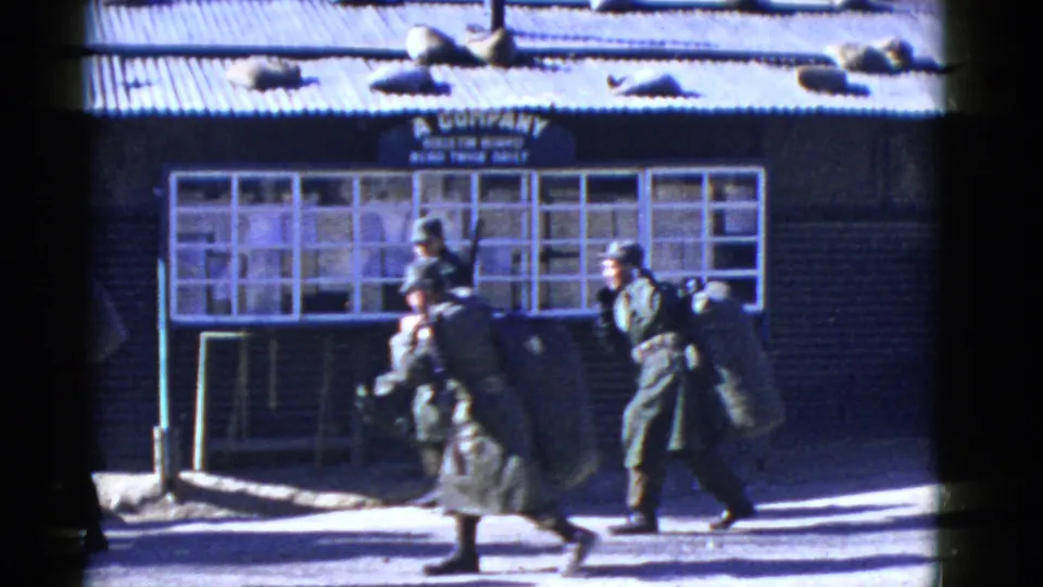Two soldiers with rifles and heavy packs move past the windowed building, while another figure is partly visible near the left edge of the frame.