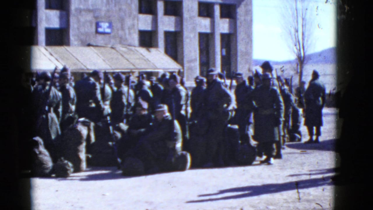 In San Francisco archival 8mm footage, uniformed men stand and sit with luggage outside a large institutional building.