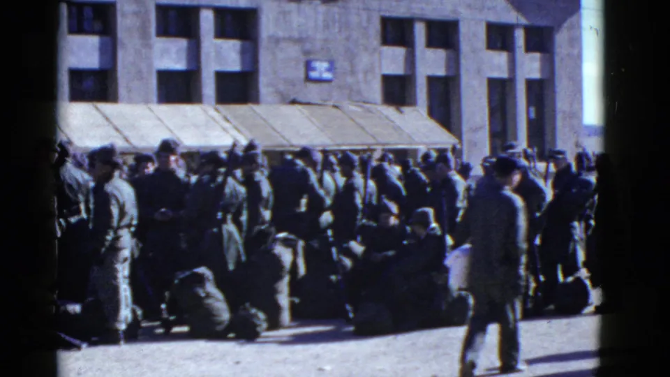 Uniformed men and a few passersby fill the sidewalk outside a large building, with bags clustered near seated figures.