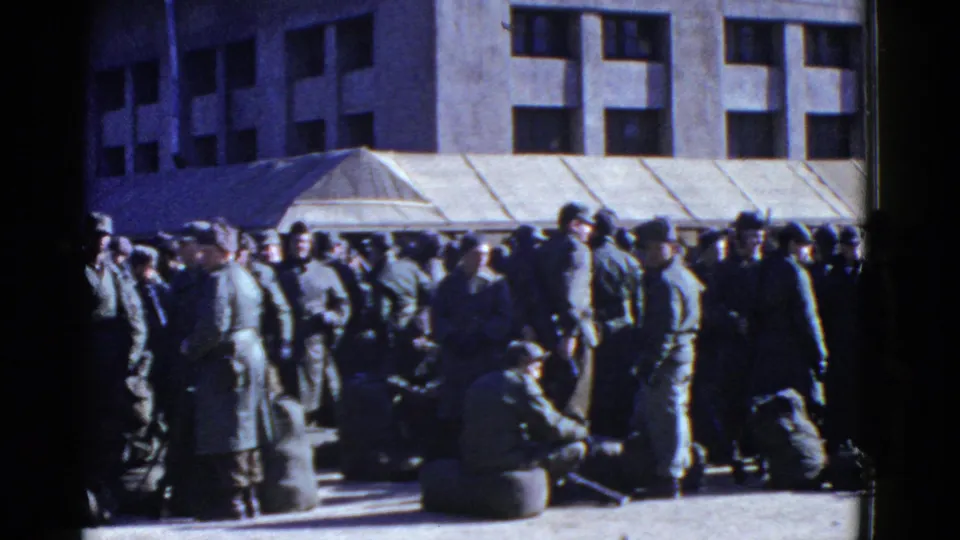 San Francisco 1950s home-movie frame showing soldiers waiting in clusters beside bags outside an institutional-looking building.