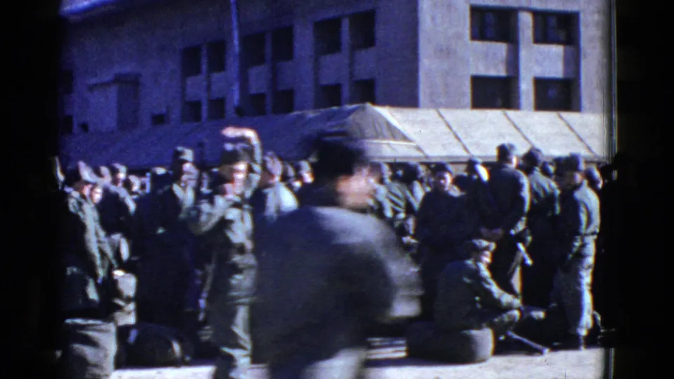 1950s San Francisco home-movie frame with motion blur as a uniformed man passes before soldiers waiting near an awning.