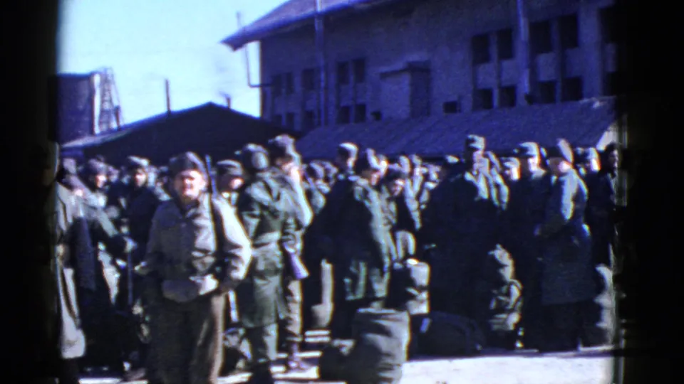 Early 1950s 8mm frame of soldiers standing outside barracks structures with bags and coats visible.