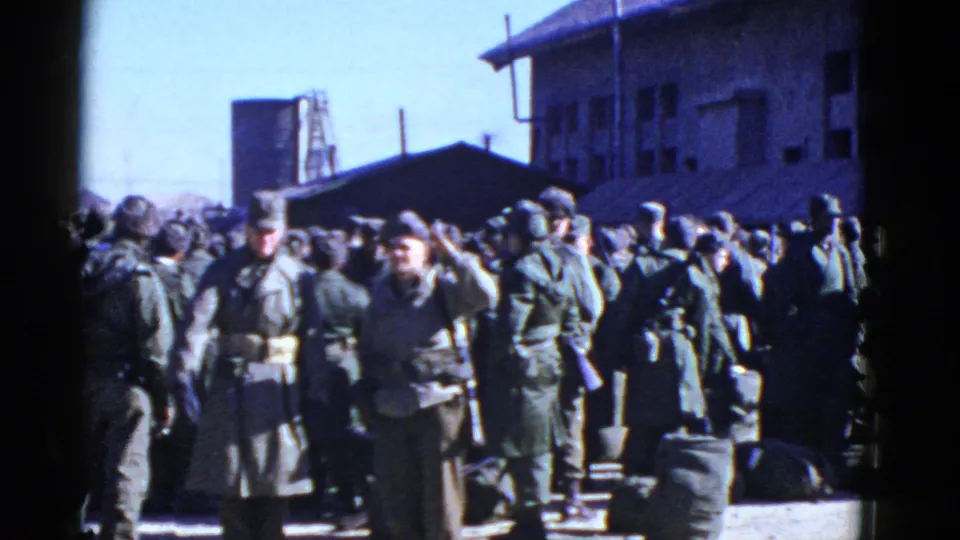 San Francisco military home-movie frame showing soldiers in olive uniforms waiting outdoors with luggage and barracks behind them.