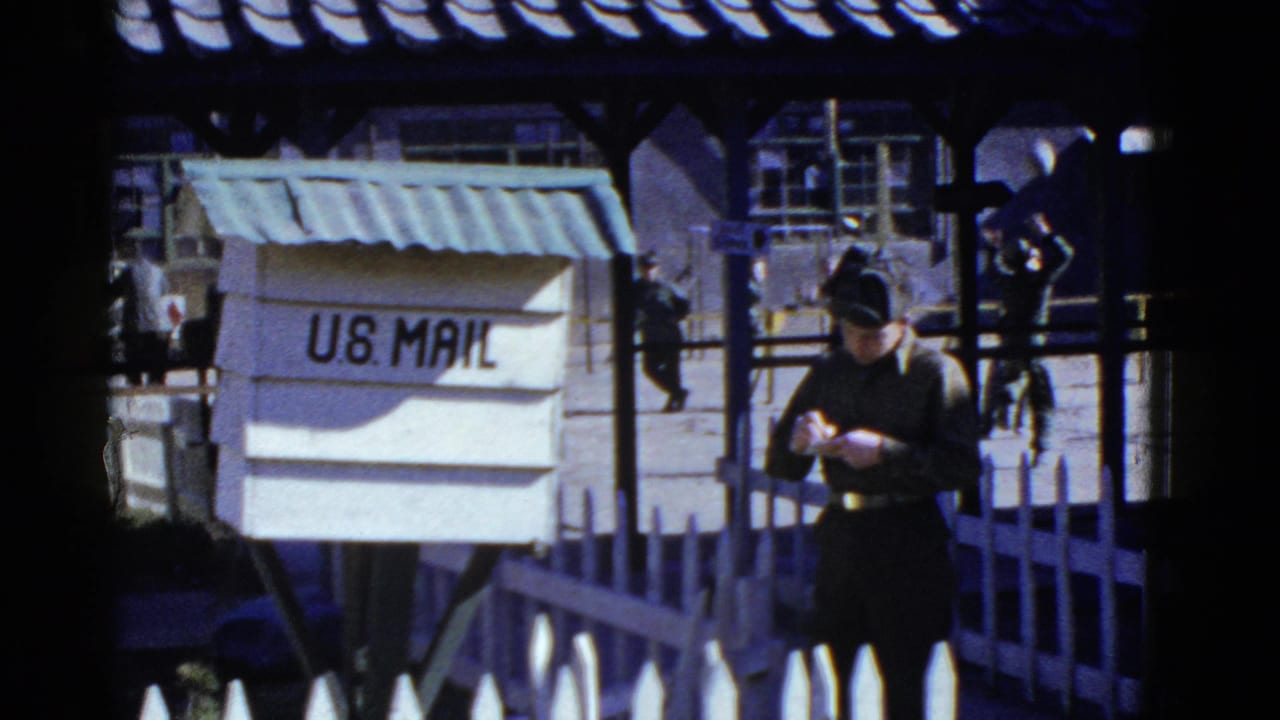 1950s San Francisco 8mm frame showing a uniformed man checking his watch beside a U.S. Mail box at a facility courtyard.