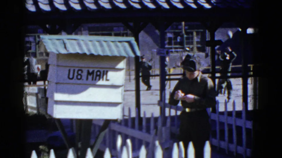 1950s San Francisco 8mm frame showing a uniformed man checking his watch beside a U.S. Mail box at a facility courtyard.