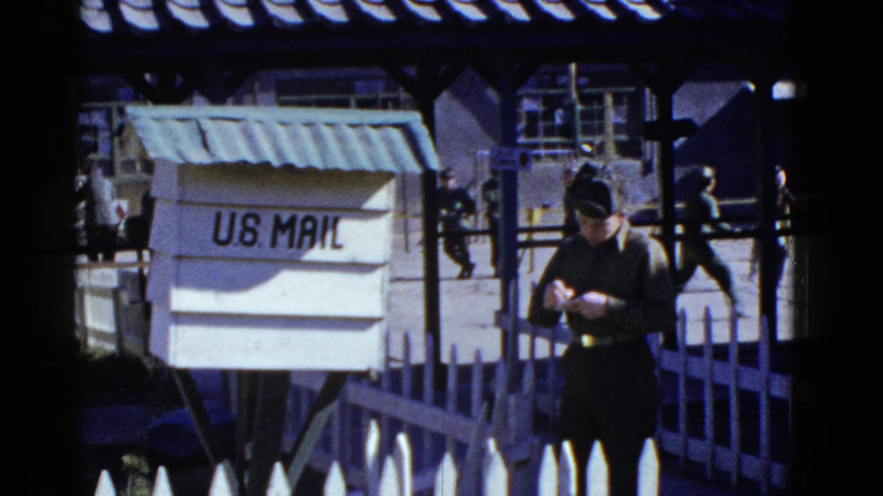 Uniformed man stands near a U.S. Mail box with other personnel in the background.