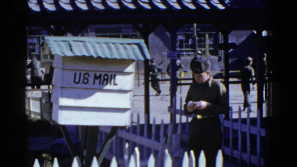 1950s San Francisco home-movie frame of a uniformed man beside a U.S. Mail box, with a courtyard and other personnel behind.