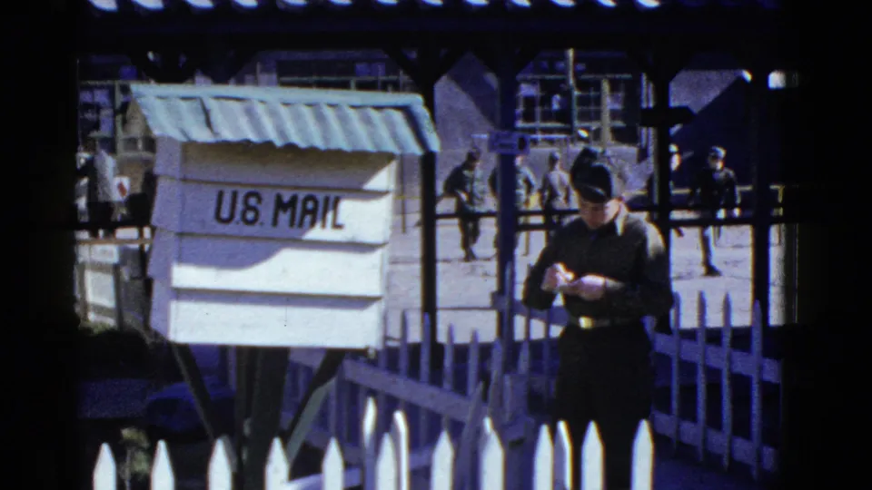 A soldier in a dark uniform appears to address or read a letter beside a U.S. Mail box, with covered base walkways behind him.