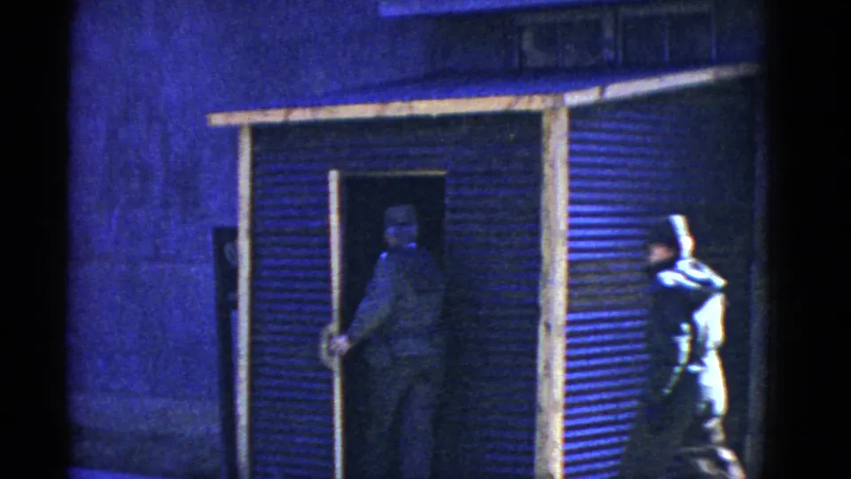A uniformed figure steps from a corrugated metal doorway as another figure stands nearby, shifting the clip into a darker blue-lit scene.