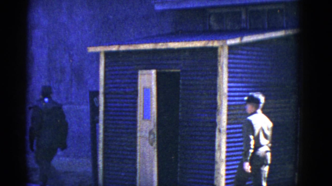 1950s San Francisco home-movie frame with two adult men near a makeshift corrugated building and open door under strong blue lighting.