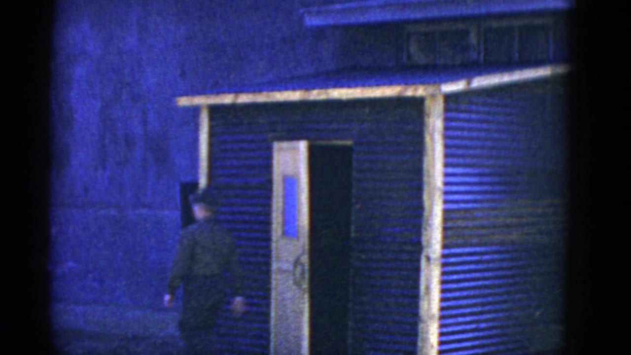 An adult man approaches an open doorway in a corrugated structure, with blue-tinted light and dark film borders visible.
