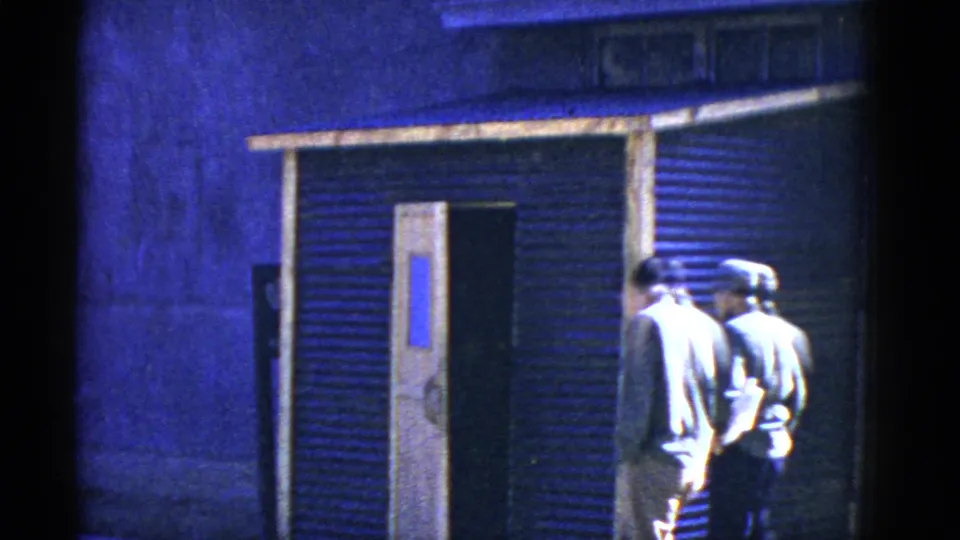 Two adult men, one wearing a cap, pass the open door of a corrugated metal building in blue-tinted 8mm footage.