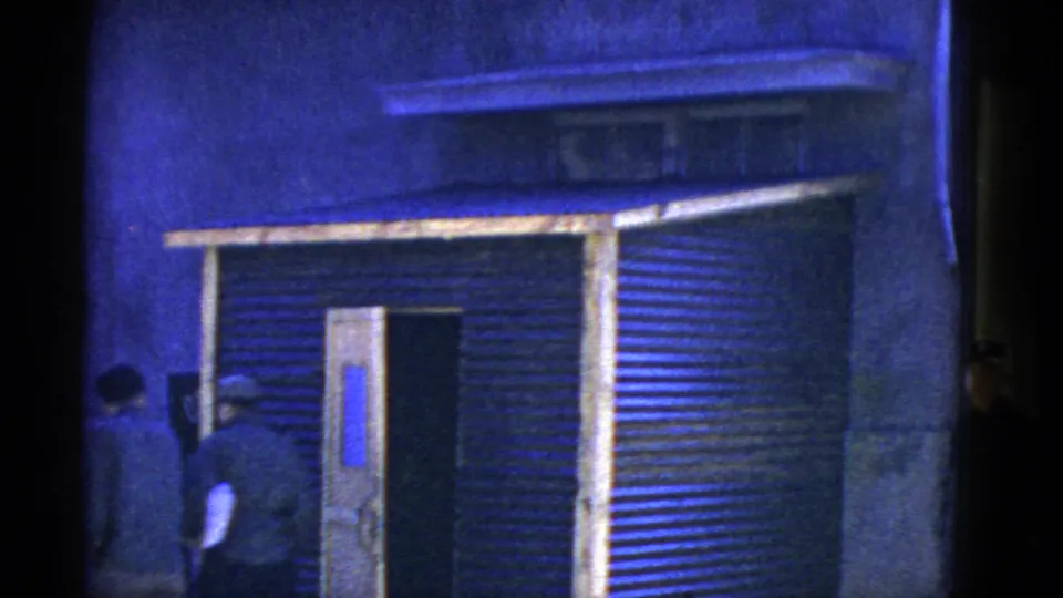 Several adult men are partially visible near a corrugated metal building and open doorway in a blue-tinted 1950s home-movie frame.