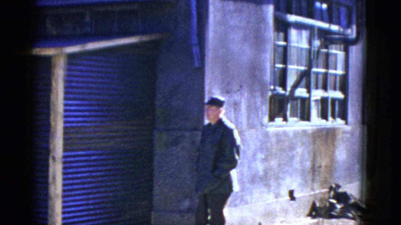 A uniformed man in a cap walks along a San Francisco exterior with a barred window and roll-up door visible behind him.