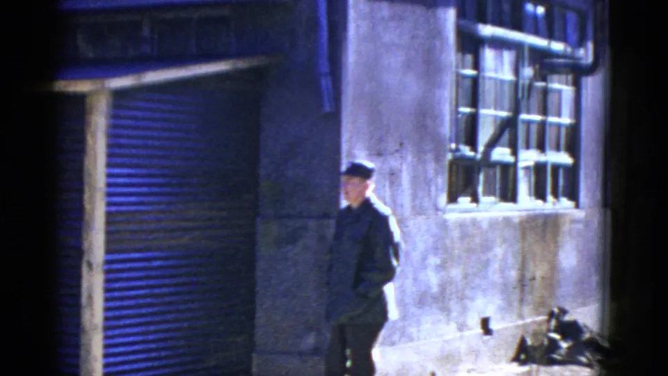 A uniformed man in a cap walks along a San Francisco exterior with a barred window and roll-up door visible behind him.