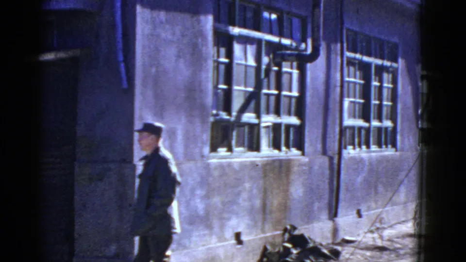 The man continues left along the exterior, with barred windows, exposed pipes, and debris visible in the alley-like setting.