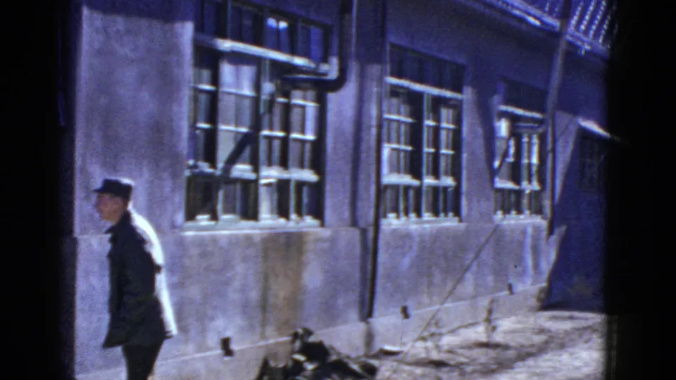 Near the end of the clip, the uniformed man reaches the left side of frame as the camera holds on barred windows and a narrow exterior passage.