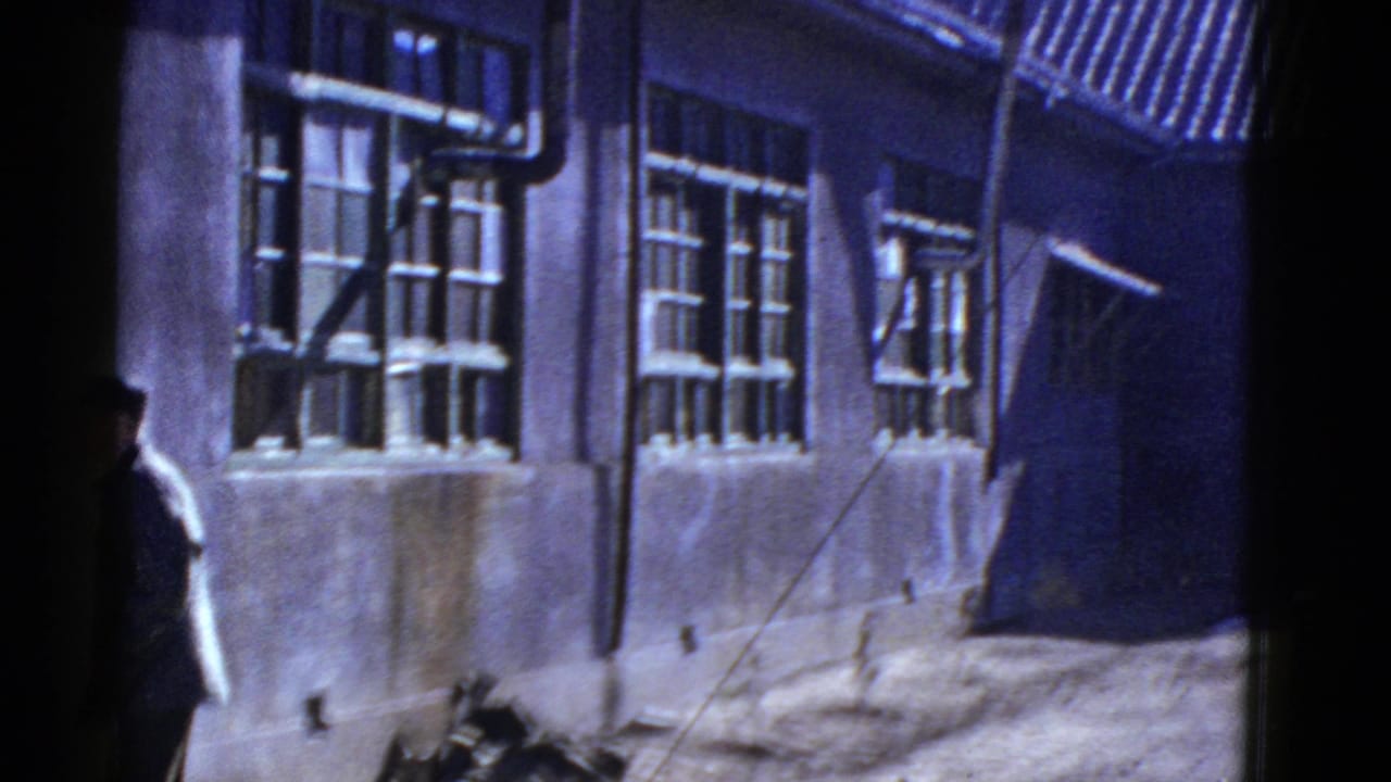 Early 1950s California home-movie frame showing a uniformed figure near a building with barred windows and exterior piping.