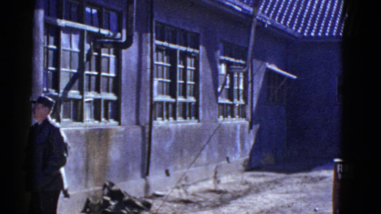 Uniformed soldier at a mid-century military or service building with barred windows, exterior pipes, and strong shadows.
