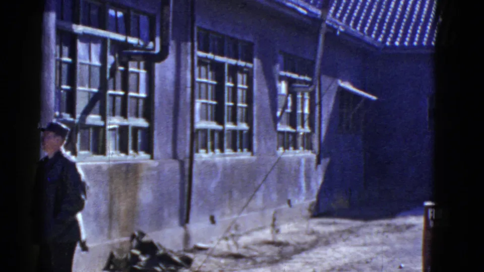 Uniformed soldier at a mid-century military or service building with barred windows, exterior pipes, and strong shadows.