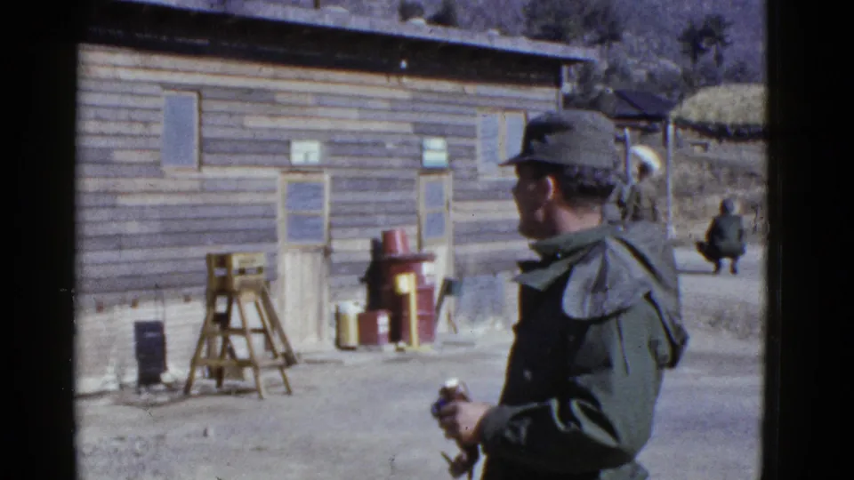 1950s home-movie view of a soldier in cap by a wooden structure with barrels, a stand, and dry hillside terrain behind.