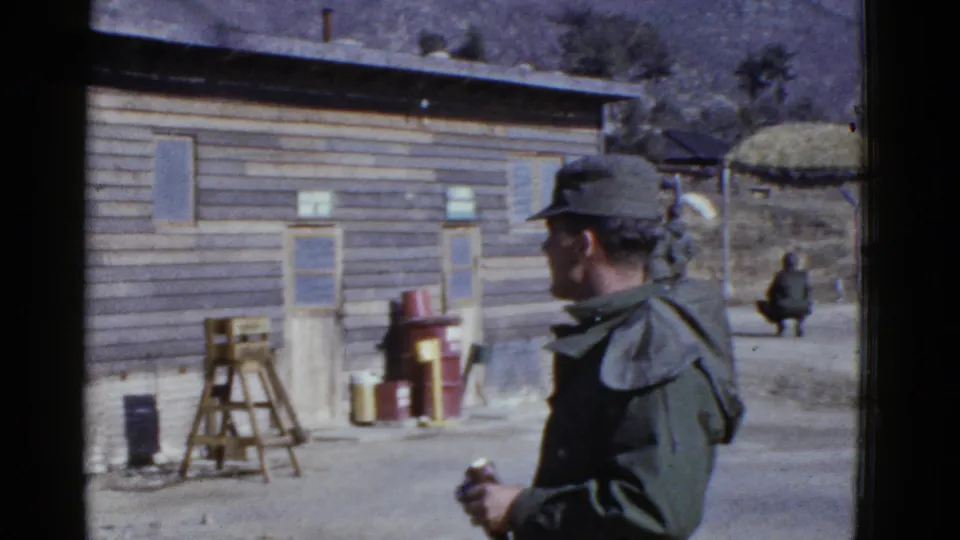 Uniformed soldier in a green jacket and cap stands near a wooden service building, with barrels and a distant figure in the yard.