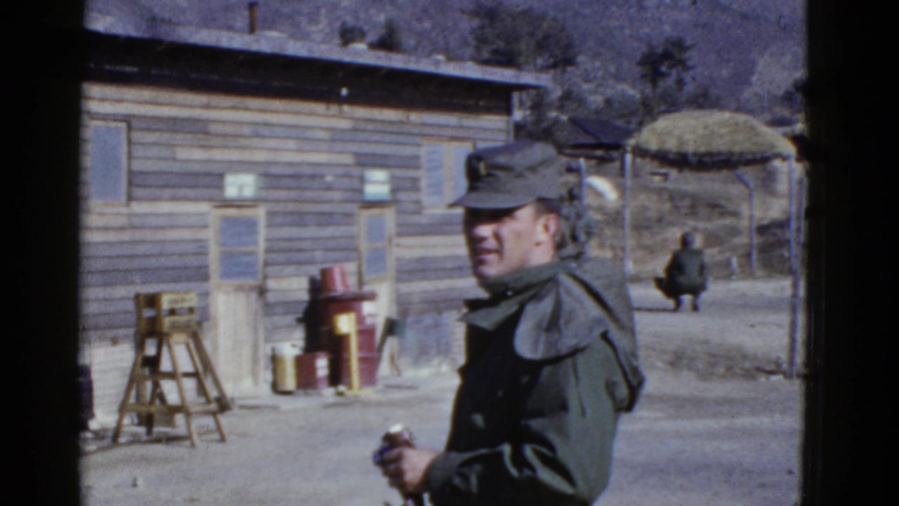 A uniformed soldier at a 1950s military camp near San Francisco stands in the foreground beside wooden buildings and camp equipment.