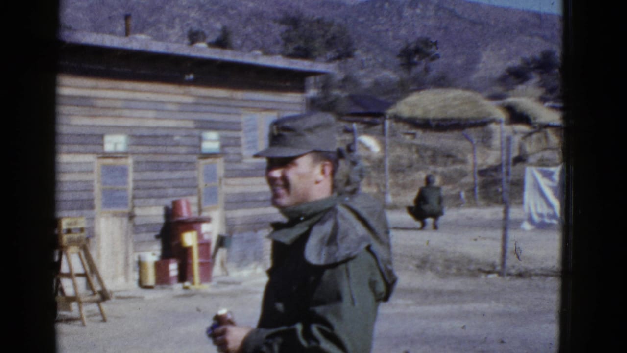 The soldier remains in the foreground holding a can, with barracks, thatched structures, and a seated figure behind him.