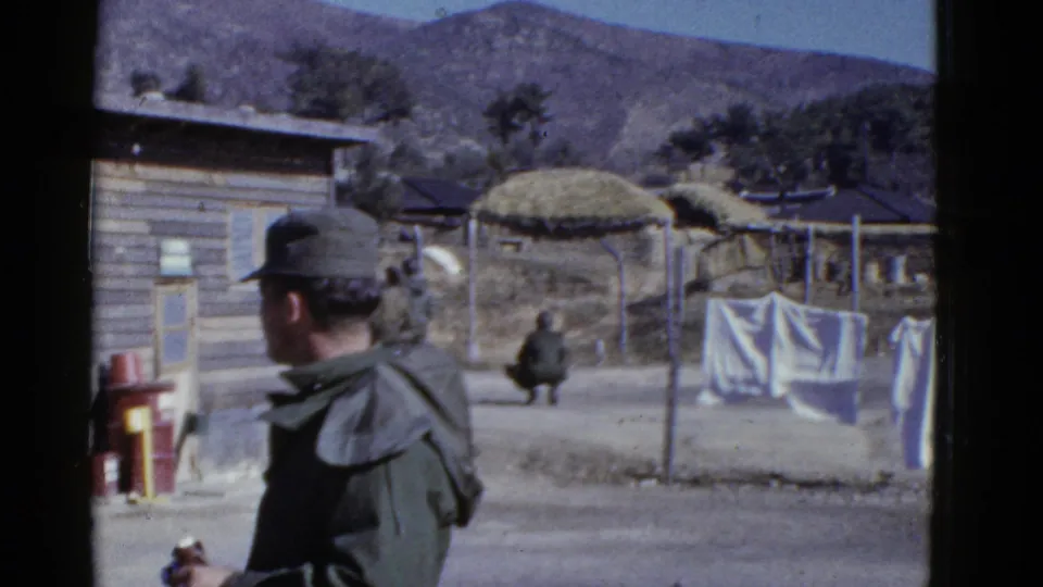 A soldier in uniform turns his back near the barracks while laundry hangs on a line and a seated figure appears across the camp yard.