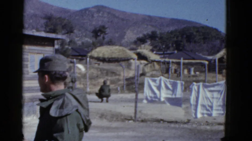 The soldier looks toward a camp area with laundry on lines, low thatched structures, fencing, trees, and hills in the background.