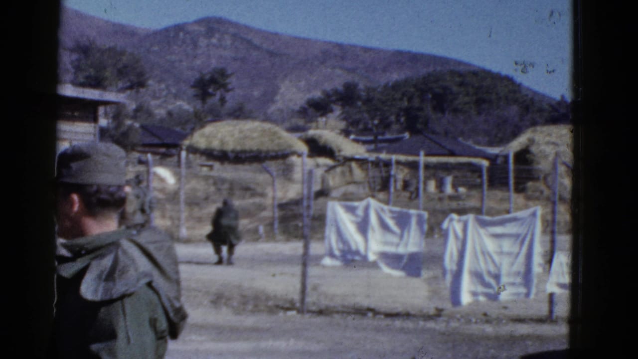 Early 1950s 8mm frame of a uniformed man looking toward fenced huts, laundry lines, and mountains in a rural village setting.