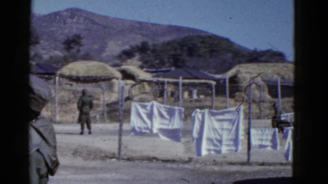 Soldier stands near hanging laundry in a fenced area with thatched huts and mountains behind.