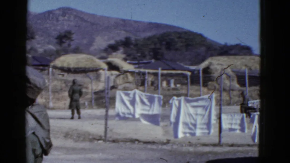 Early 1950s rural village view with a soldier near laundry lines, thatched structures, fencing, and a mountainous backdrop.