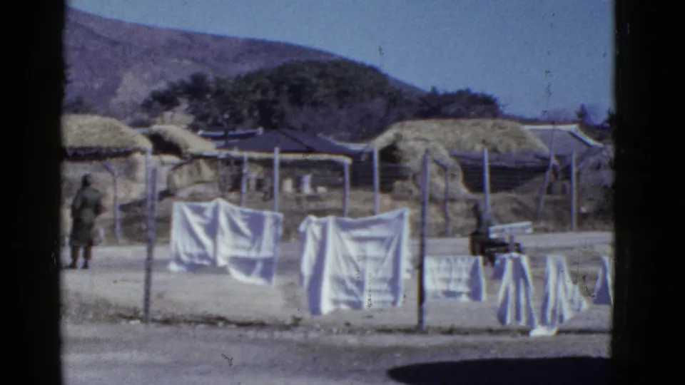 A uniformed figure moves through a dusty village area with hanging laundry, thatched roofs, fencing, and hills in the background.